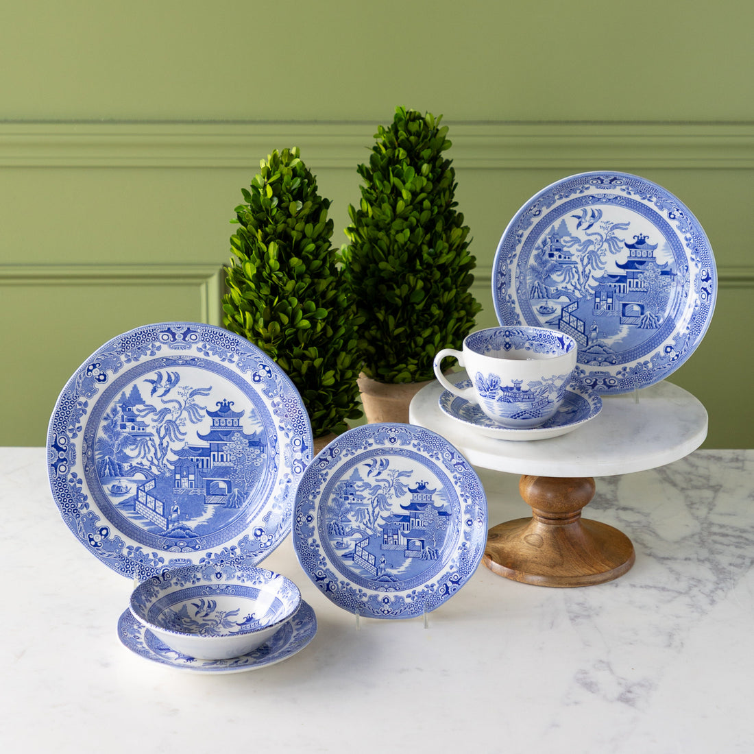 Burleigh Blue Willow Dinnerware displayed on a marble table with topiary plants behind it.