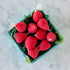 A blue carton filled with ten red, strawberry-shaped candles made from pure beeswax, on green paper grass, set on a white marble surface.