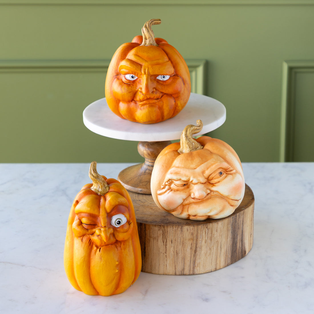 Three carved pumpkins with facial expressions on a marble surface with a green wall background.