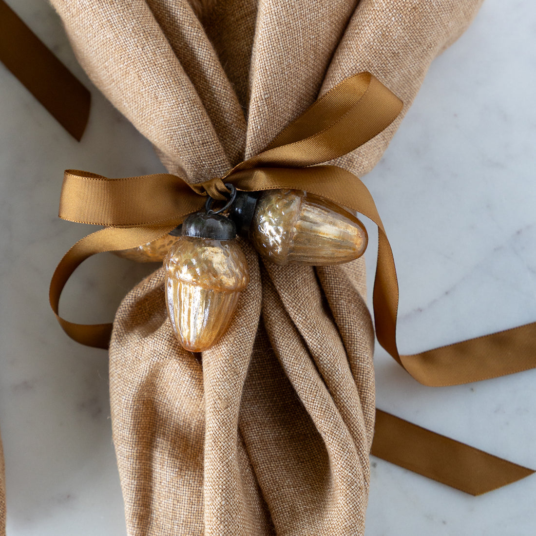 A brown napkin tied together with a brown silk ribbon and two Mercury Glass Acorn Shaped Ornaments.