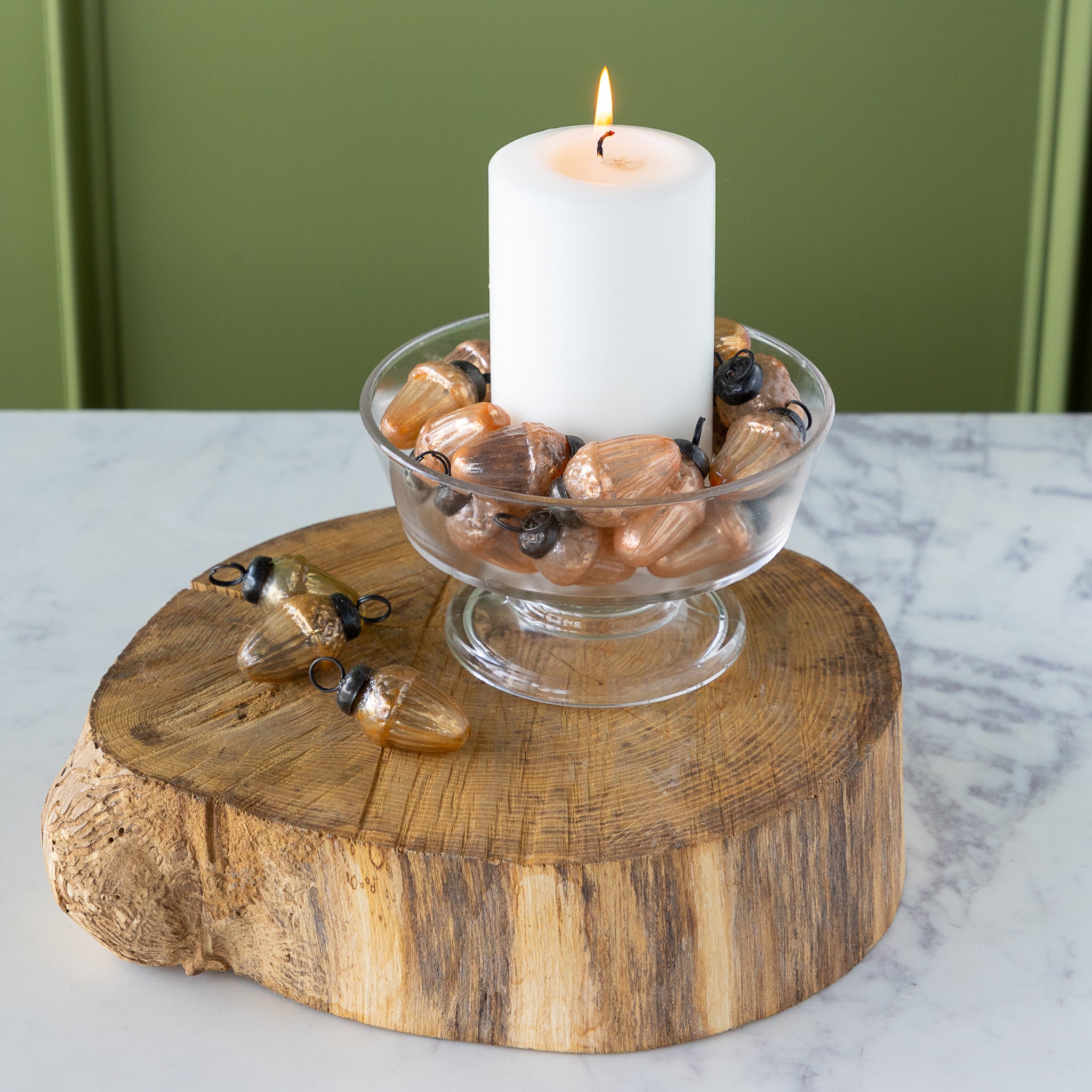 Candle in a glass holder filled with  Mercury Glass Acorn Shaped Ornaments on a wooden base, against a green wall.