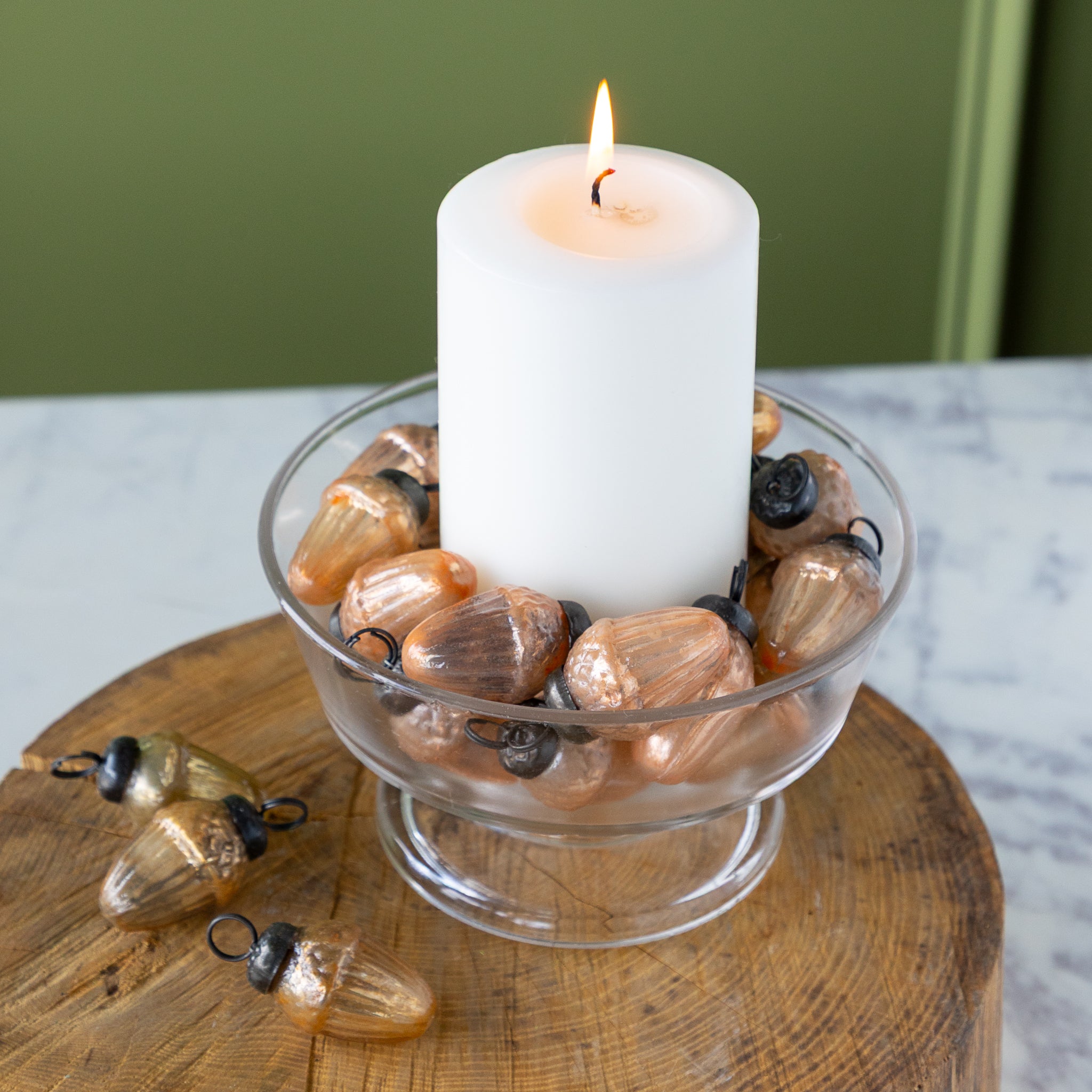 Candle in a glass holder filled with  Mercury Glass Acorn Shaped Ornaments on a wooden base, against a green wall.