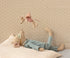 Stuffed animal toy lying on a bed with another toy hanging above against a patterned wall.
