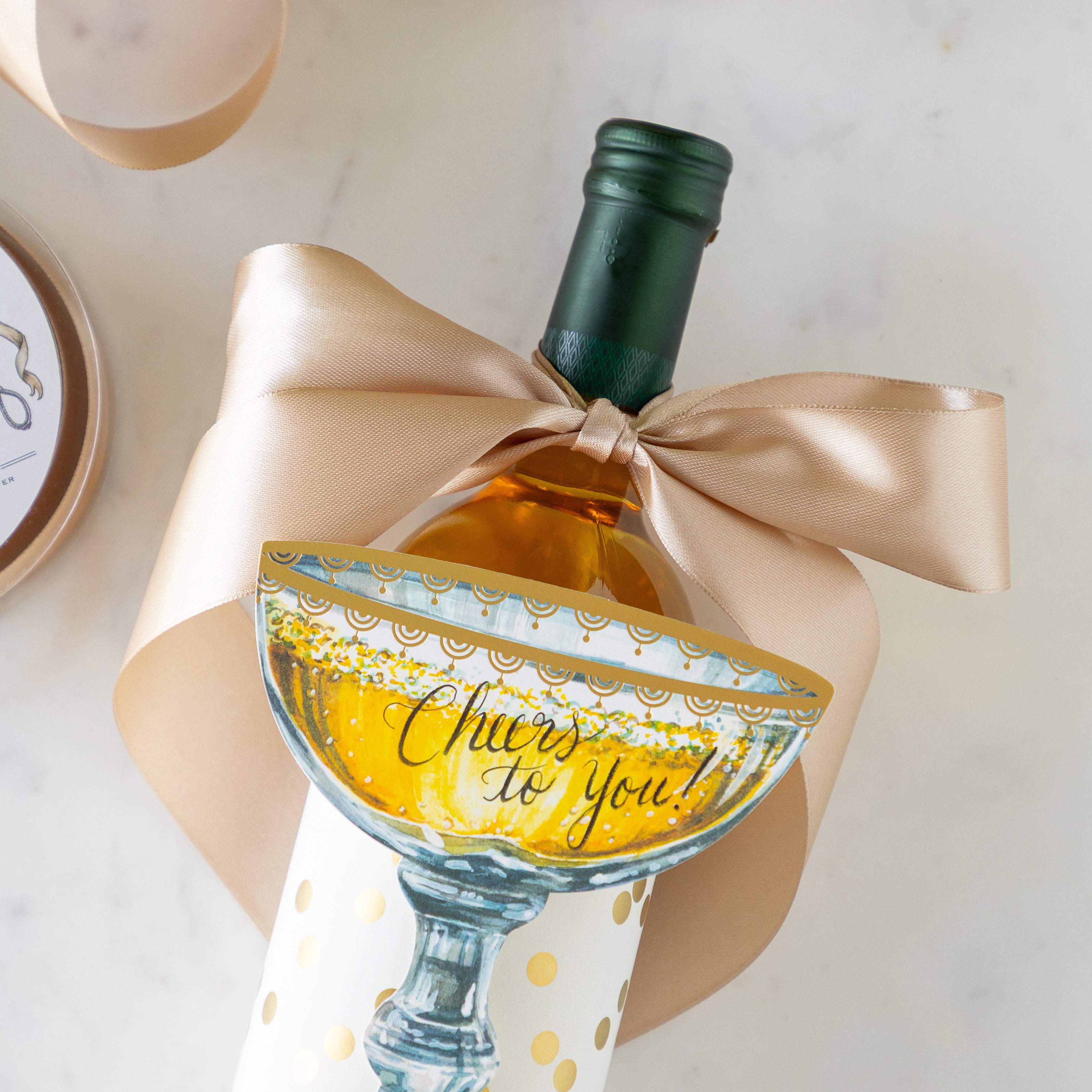 A bottle of wine with the Fancy Champagne Ribbon tied in a bow around it, wrapped in the Gold Confetti Placemat and Champagne Coupe Place Card atop reading &quot;Cheers to You!&quot;.