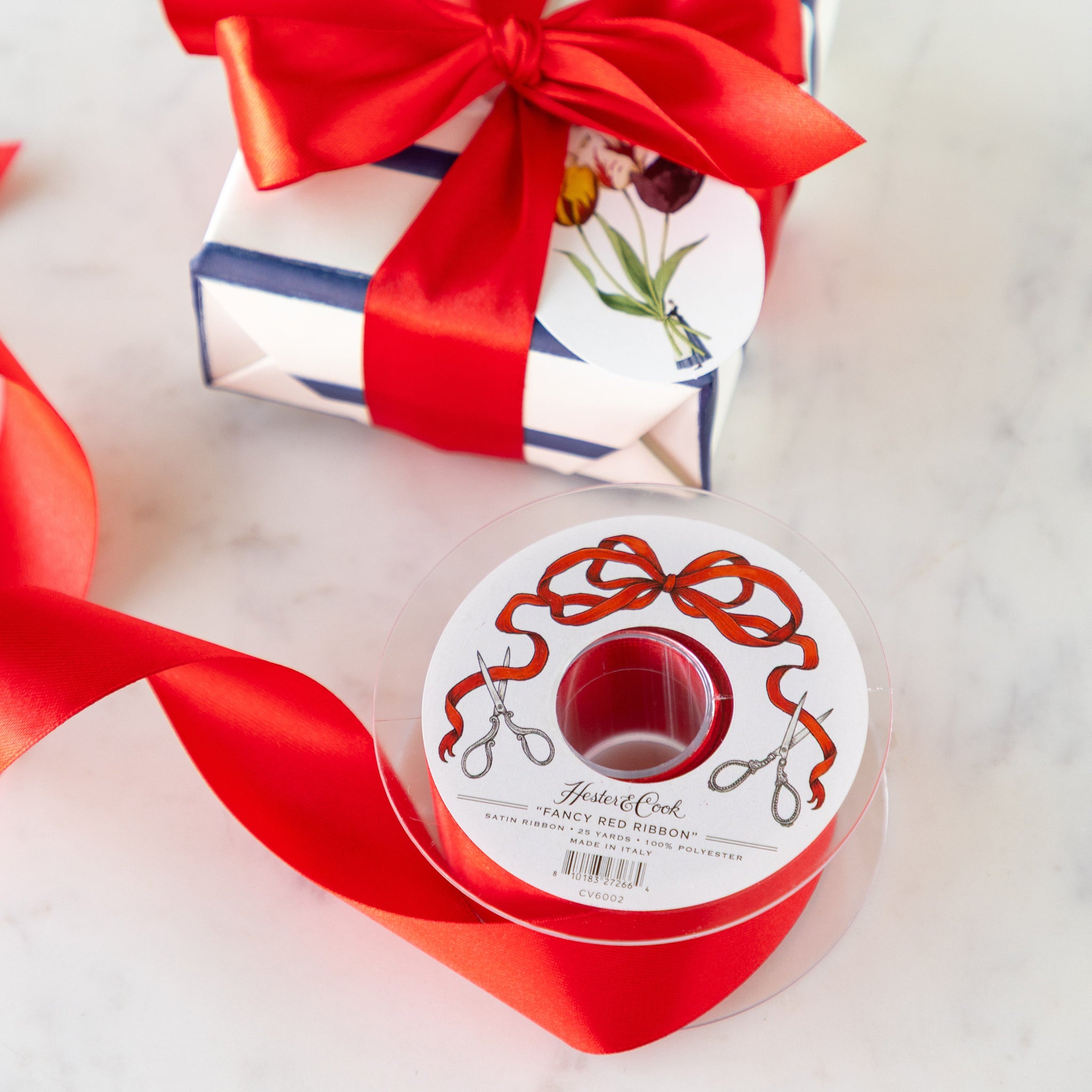 Spool of Fancy Red Ribbon with a decorative label next to a wrapped gift with a red ribbon bow.