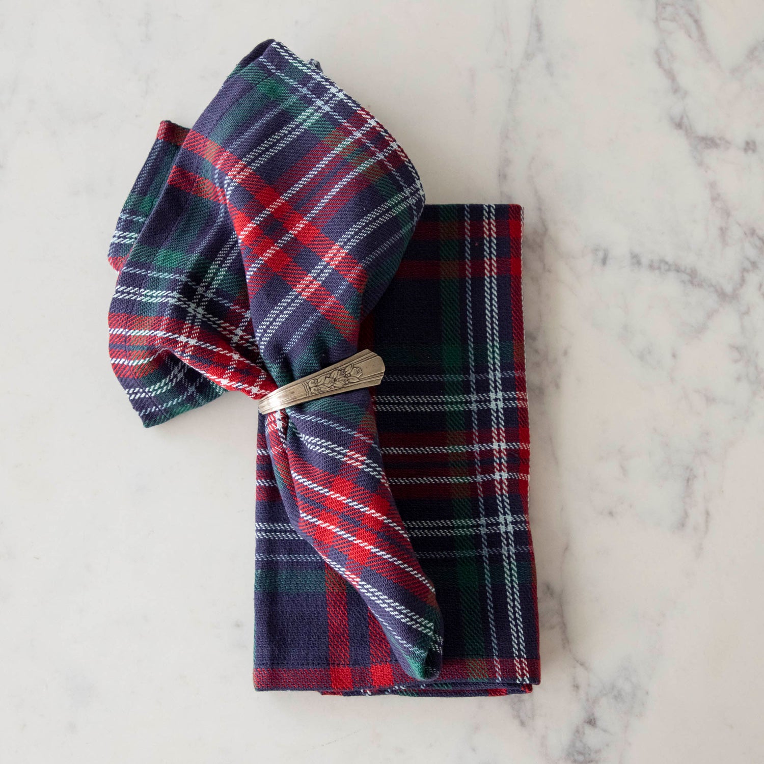 Two blue and red plaid cotton napkins, one folded and one in a napkin ring, displayed on a white surface.