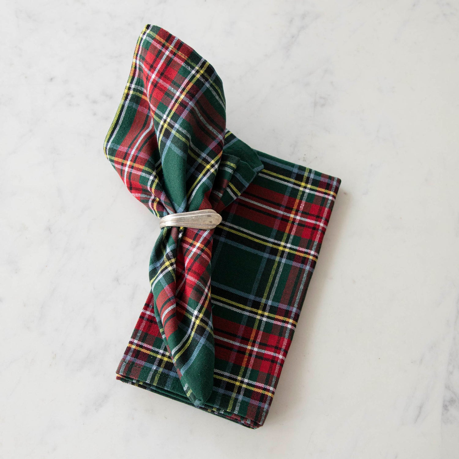 Two green and red plaid cotton napkins, one folded and one in a napkin ring, displayed on a white surface.