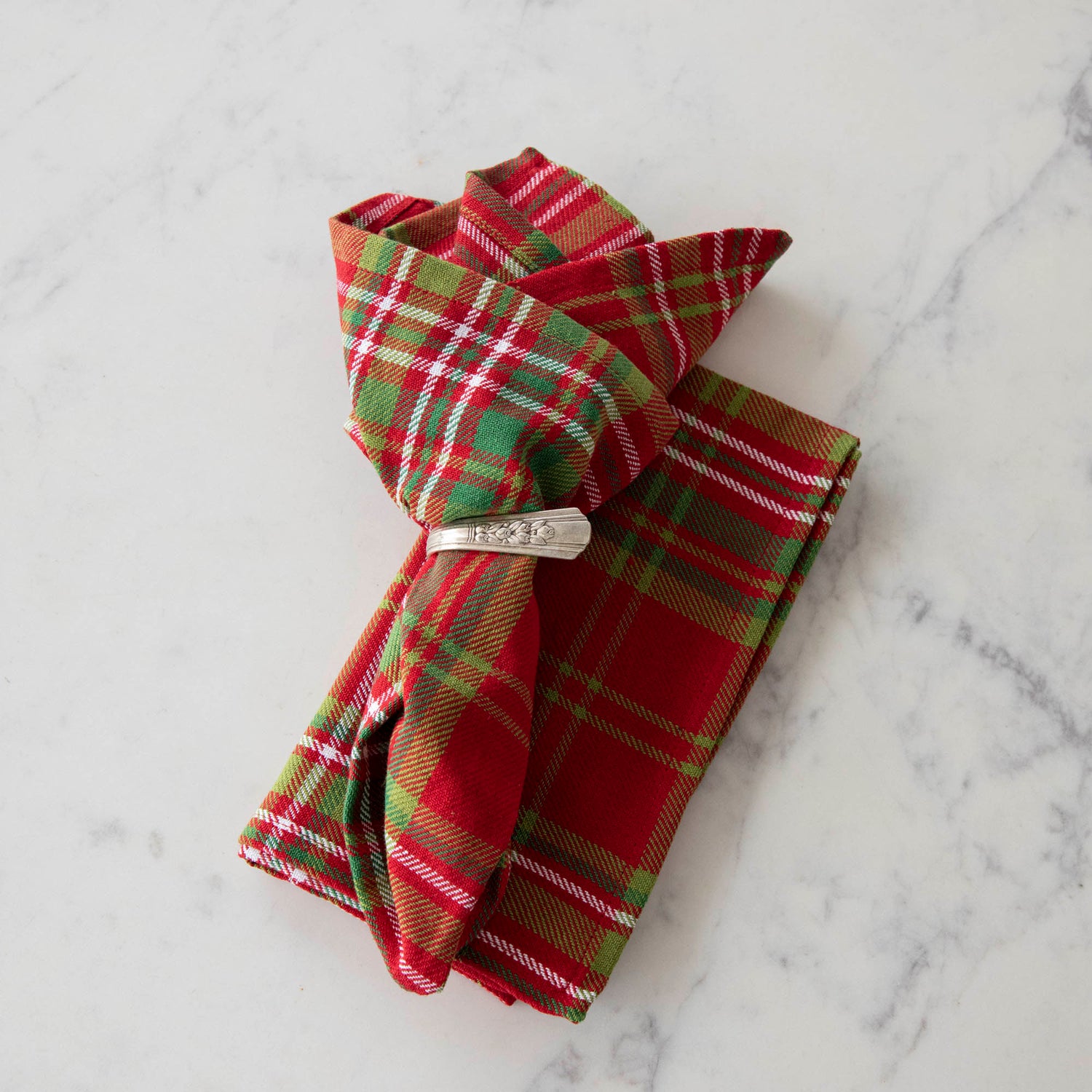 Two red and green plaid cotton napkins, one folded and one in a napkin ring, displayed on a white surface.