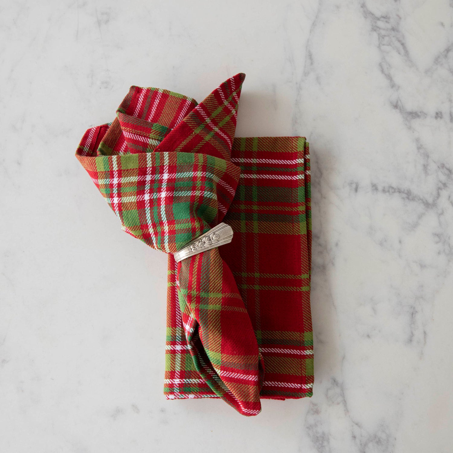 Two red and green plaid cotton napkins, one folded and one in a napkin ring, displayed on a white surface.