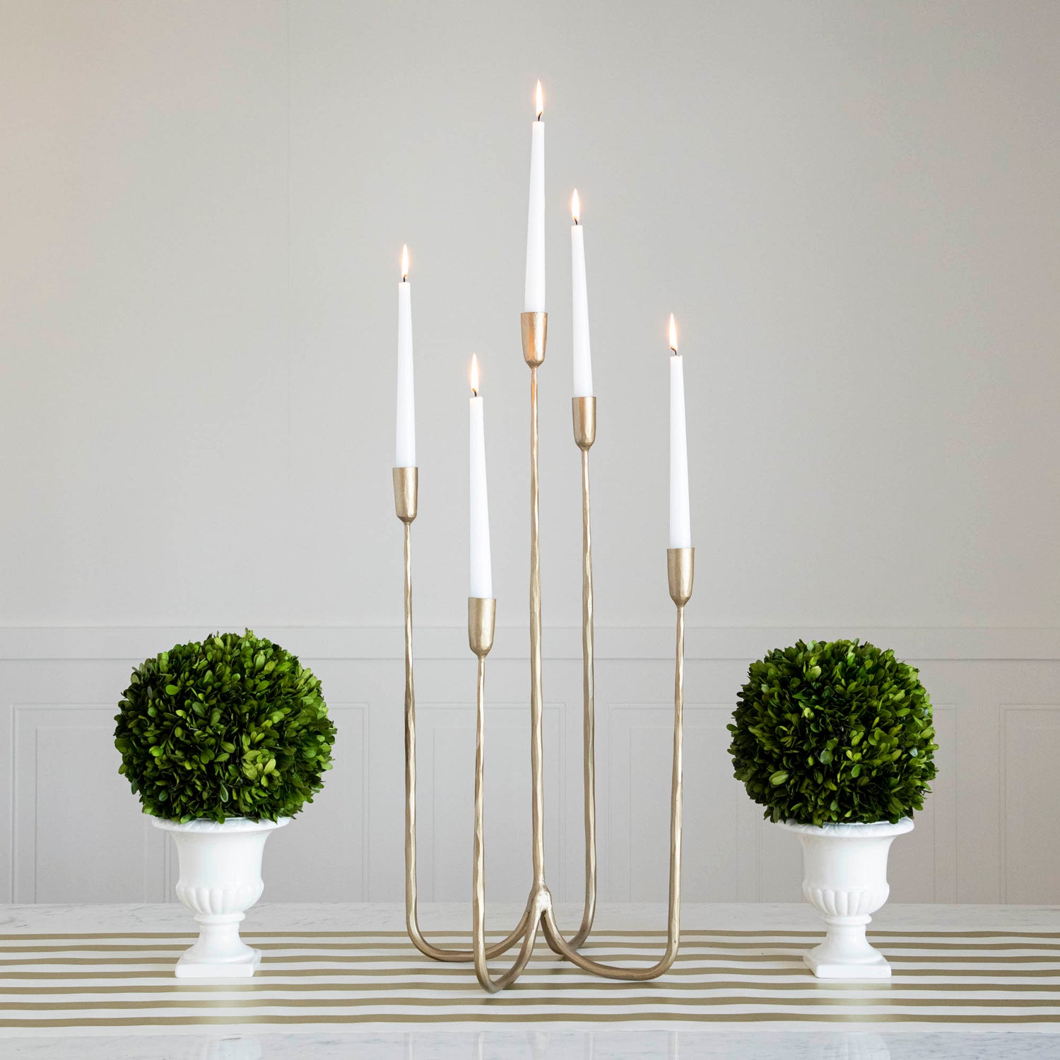 Forged Brass Candelabra in tall size with white candles lit in it on a table with vases that have boxwood balls on them. 