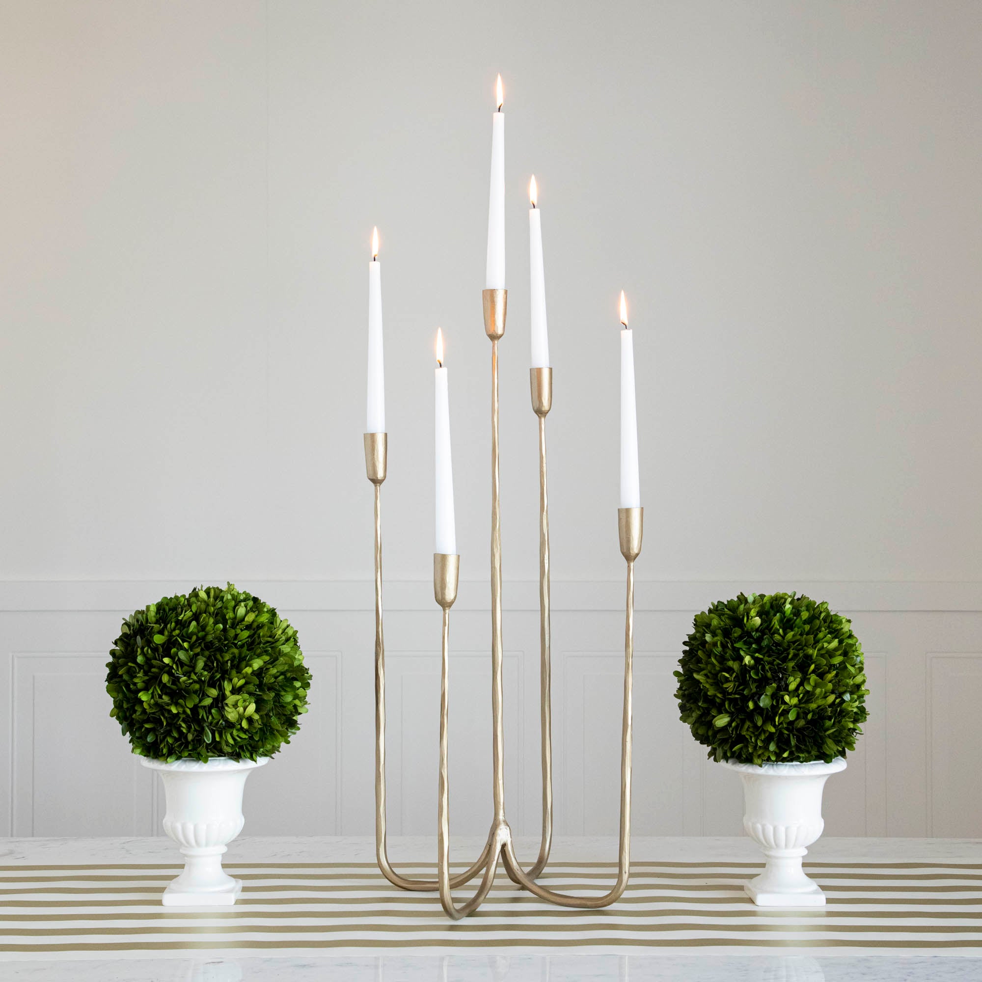 Forged Brass Candelabra in tall size with white candles lit in it on a table with vases that have boxwood balls on them. 