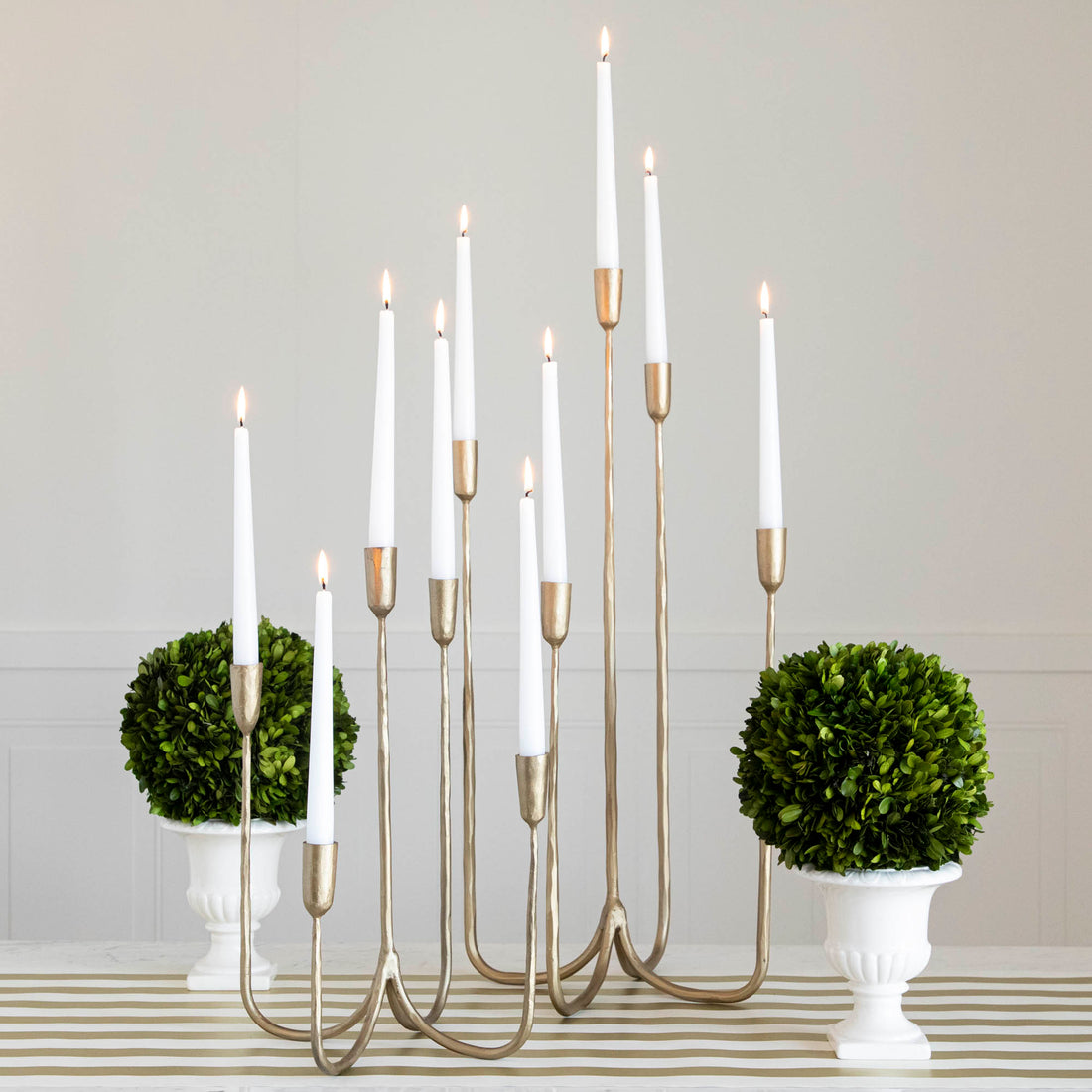 Forged Brass Candelabra in short and tall sizes with white candles lit in them on a table with vases that have boxwood balls on them. 