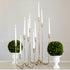 Forged Brass Candelabra in short and tall sizes with white candles lit in them on a table with vases that have boxwood balls on them. 