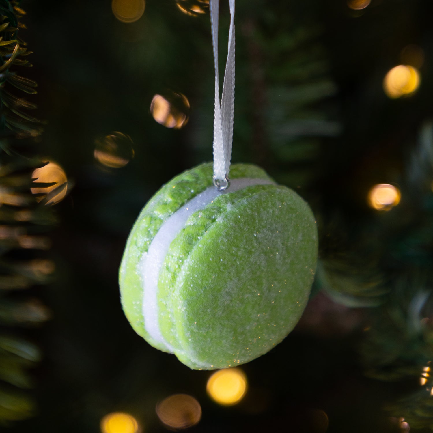 Green macaron ornament hanging on a Christmas tree with blurred lights in the background.