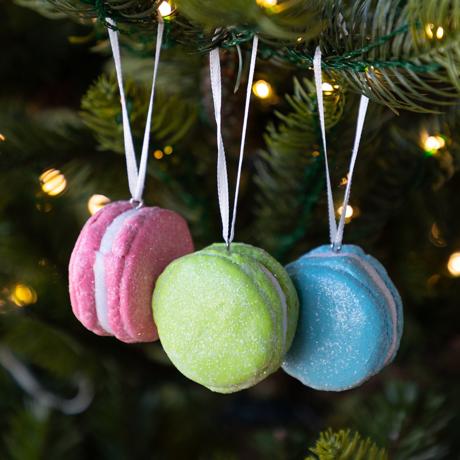 Three macaron-shaped ornaments in blue, green, and pink hanging on a Christmas tree.