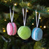 Three macaron-shaped ornaments in blue, green, and pink hanging on a Christmas tree.
