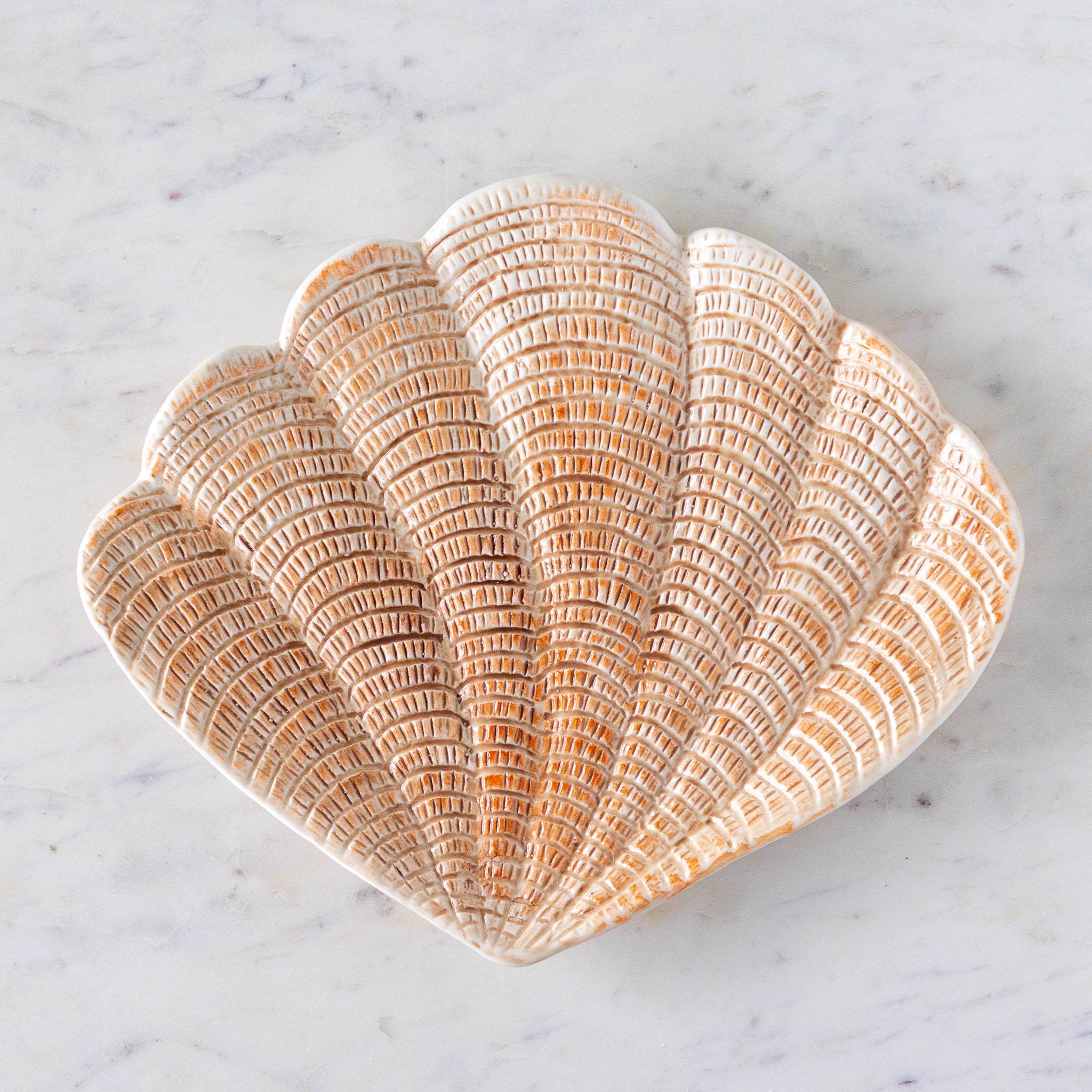 A hand-painted stoneware dish shaped like a scallop shell with textured, light peach detailing, placed on a white marble surface.