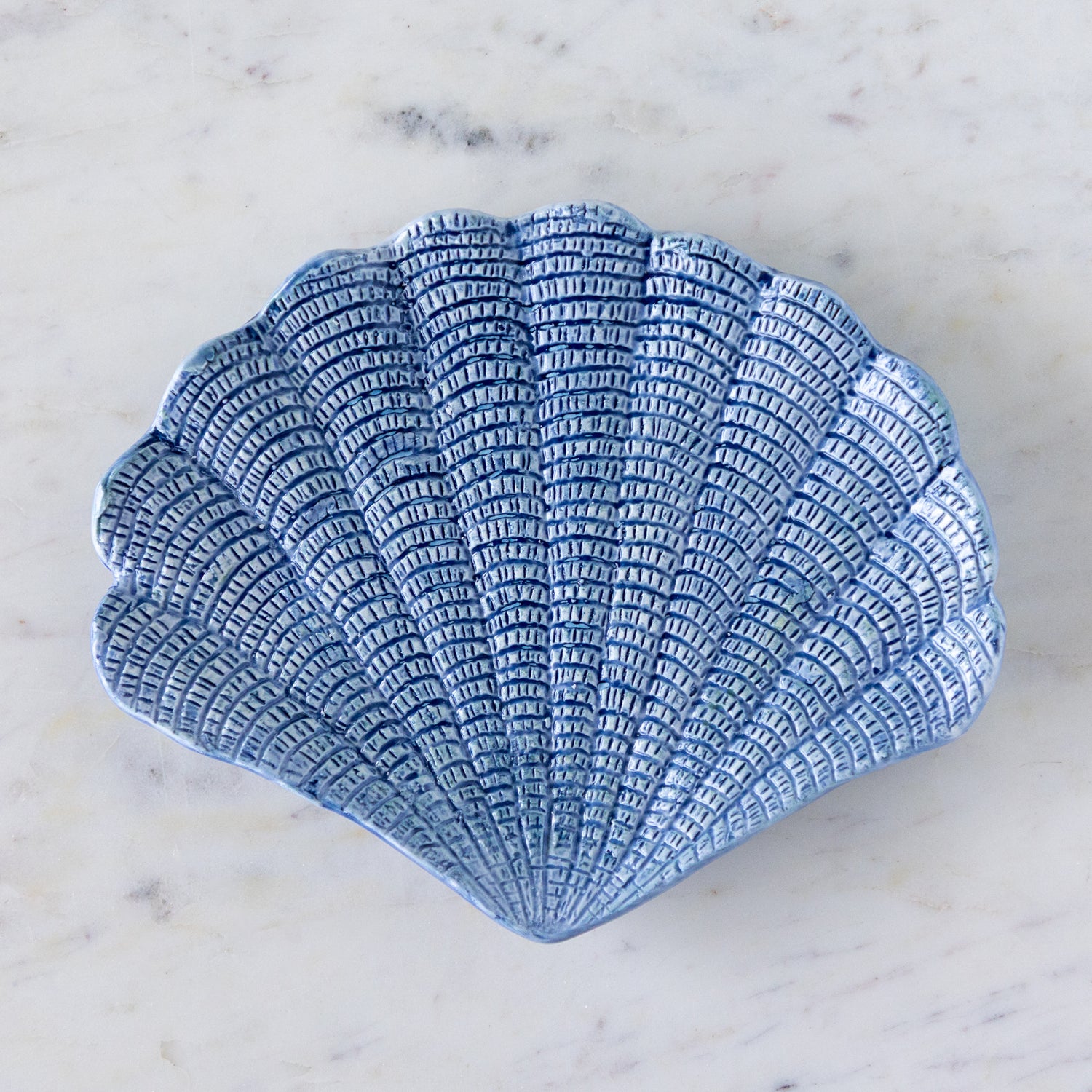 A hand-painted stoneware dish shaped like a scallop shell with textured, blue detailing, placed on a white marble surface.