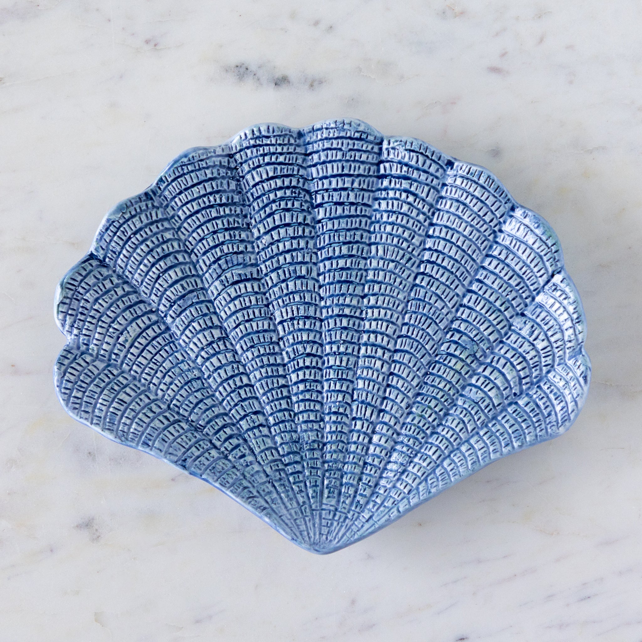 A hand-painted stoneware dish shaped like a scallop shell with textured, blue detailing, placed on a white marble surface.