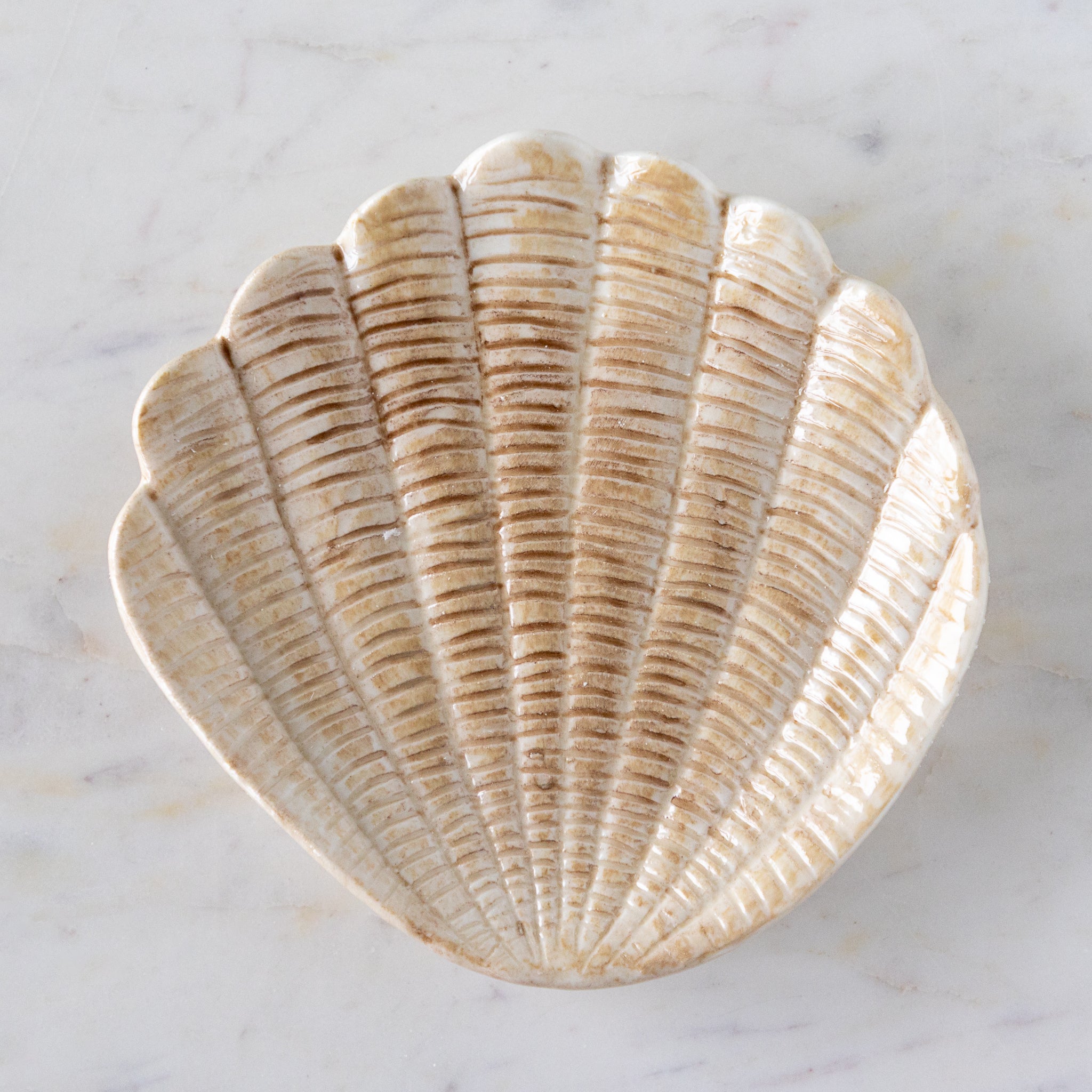 A hand-painted stoneware dish shaped like a scallop shell with textured, light tan detailing, placed on a white marble surface.