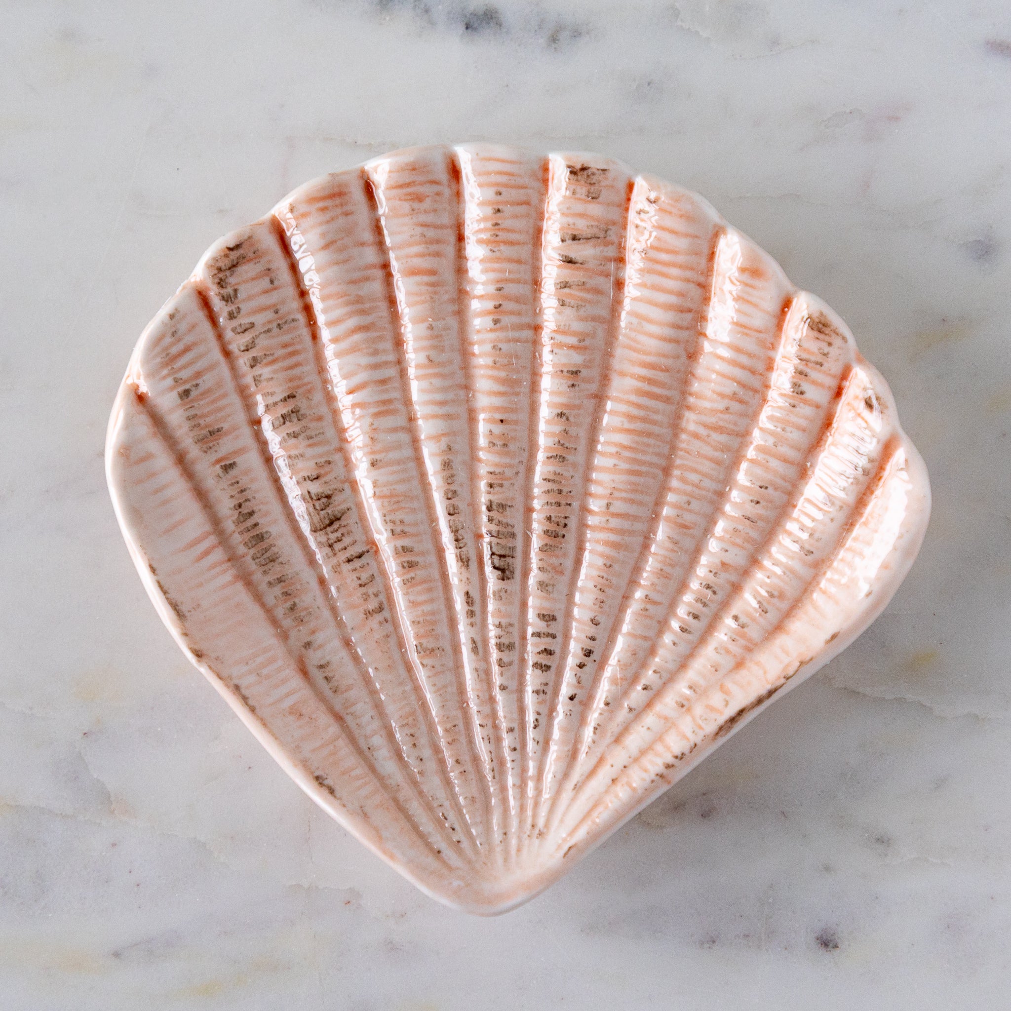 A hand-painted stoneware dish shaped like a scallop shell with textured, light pink detailing, placed on a white marble surface.