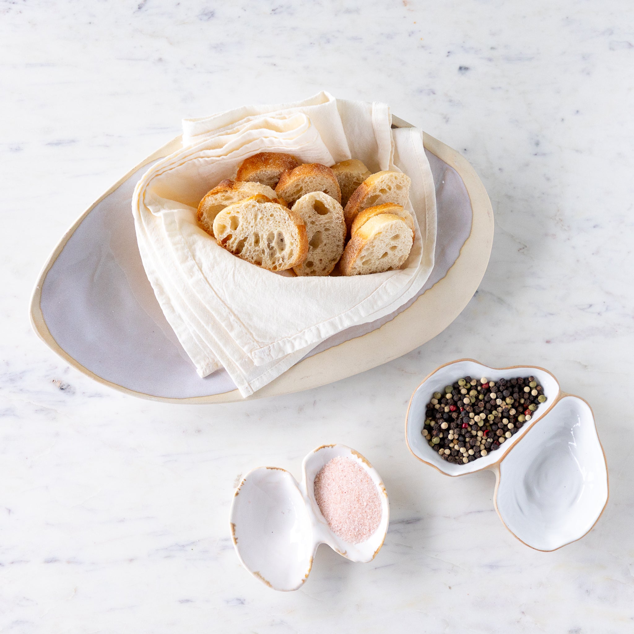 Sliced baguette on a cloth in the Single Large Oyster Shell Dish, pink salt in the Double Shell and peppercorns in the Oyster Shell.