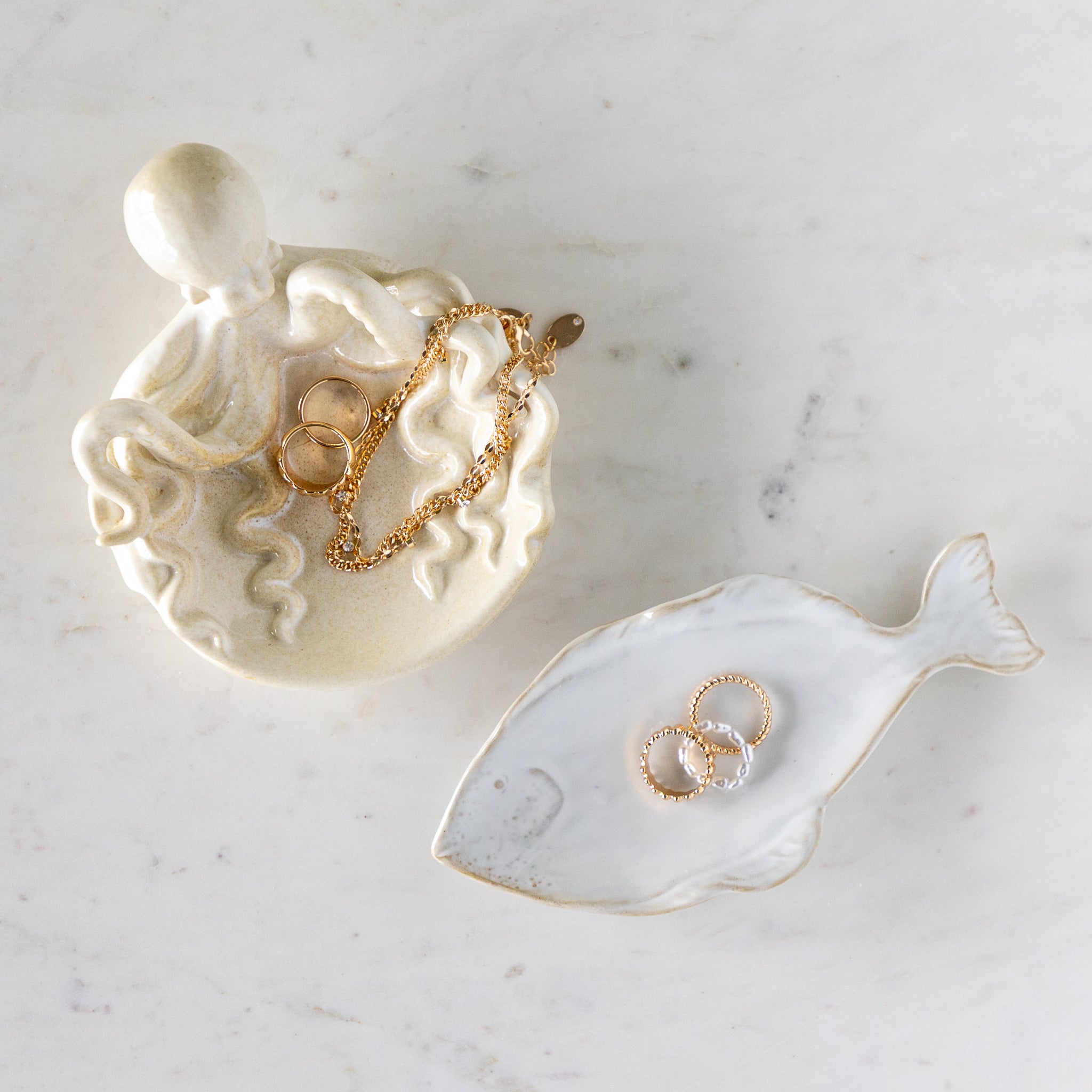 Stoneware Fish Shaped Plate with rings in it, next to Stoneware Octopus Dish filled with rings and a necklace, on a marble table.