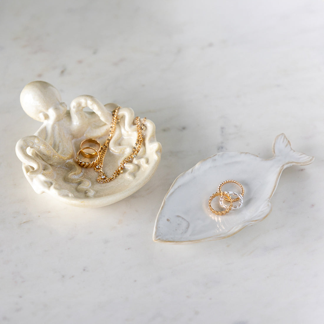 Stoneware Fish Shaped Plate with rings in it, next to Stoneware Octopus Dish filled with rings and a necklace, on a marble table.