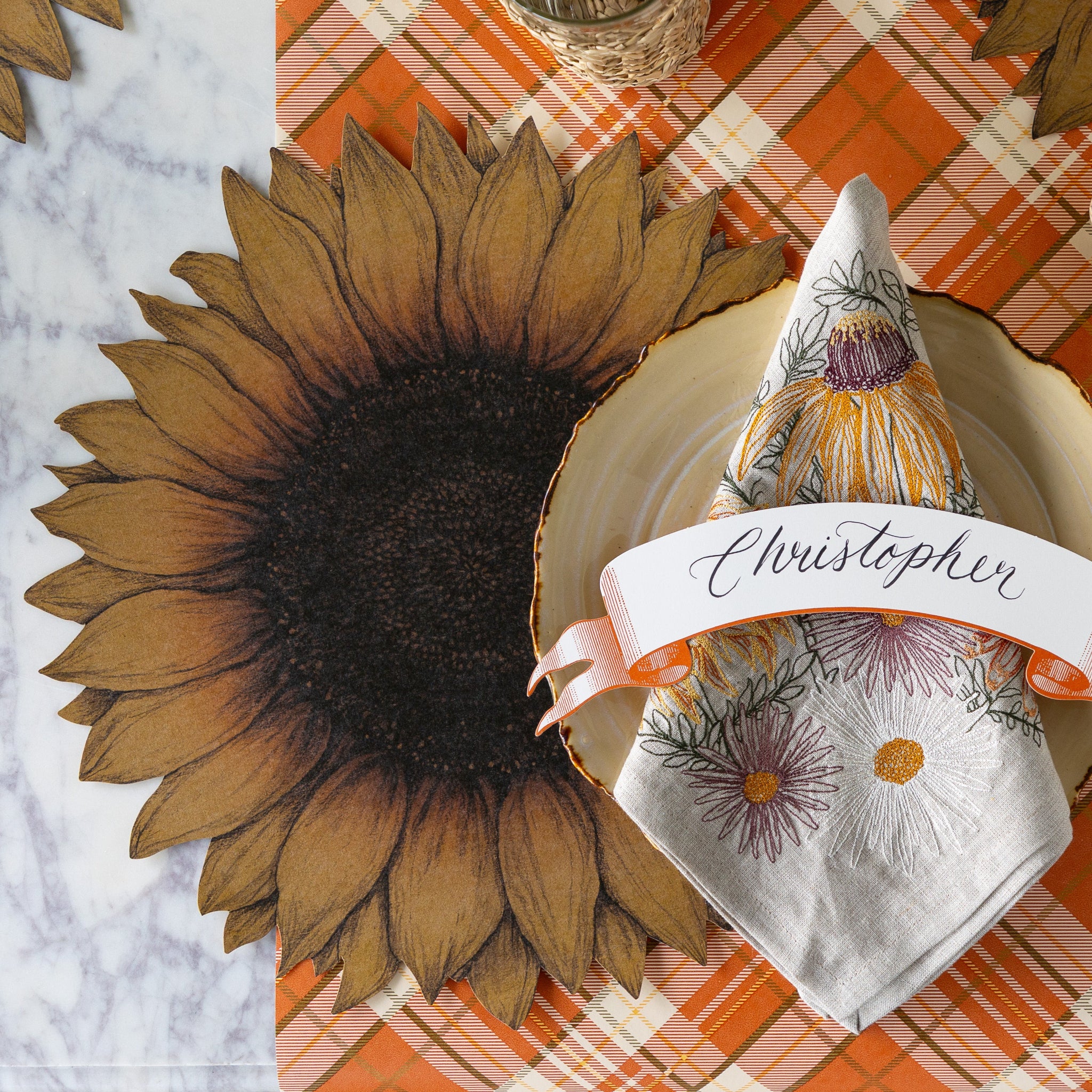 A fall place setting with the Die-cut Vintage Sunflower Placemat, wood slice plate with the Autumn Mums Napkin and Classic Orange Banner Table Accent atop, on the Orange Plaid Runner.