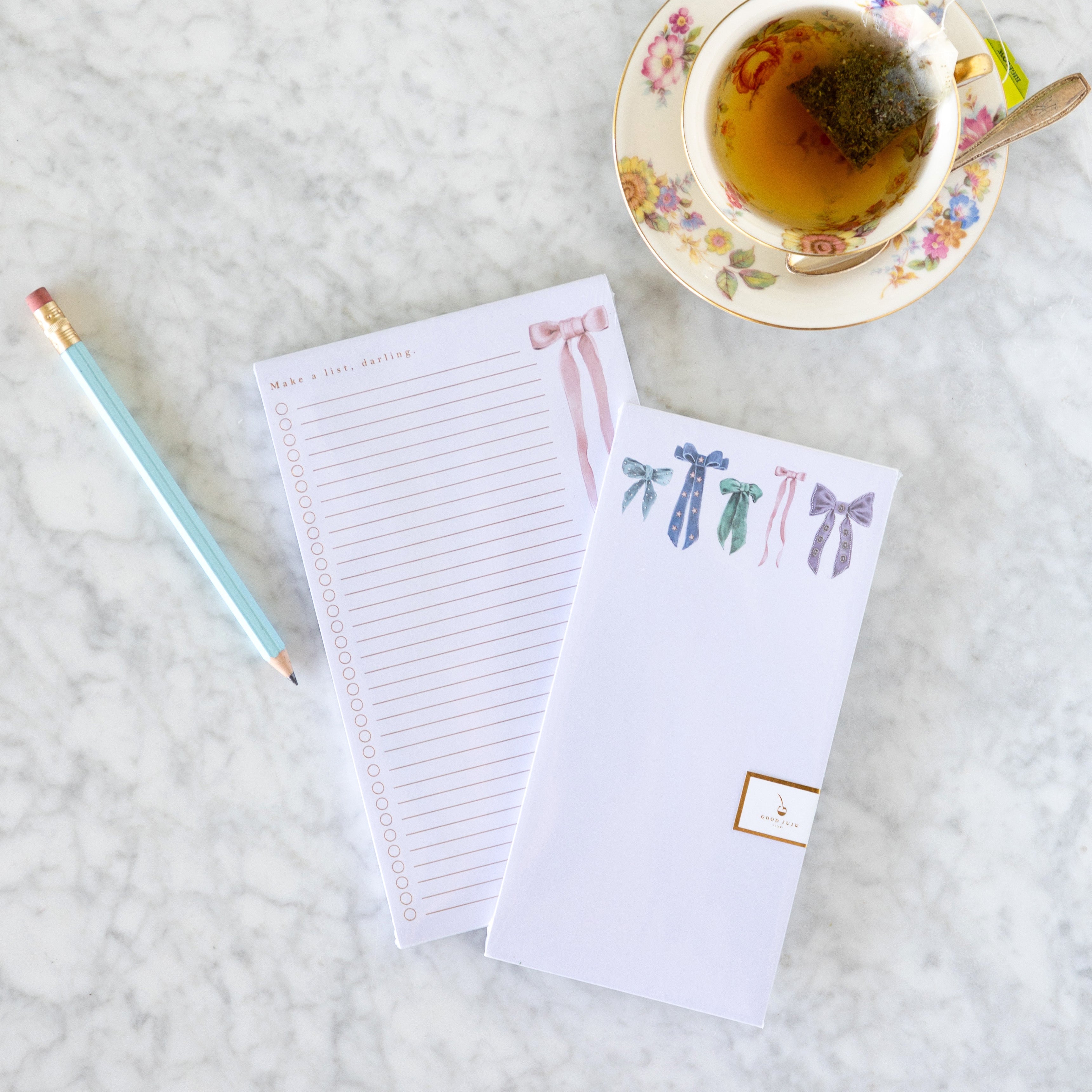 Assorted Antique Bows Notepad over top of the Make A List Darling Large Notepad with a pencil and cup of tea next to it.