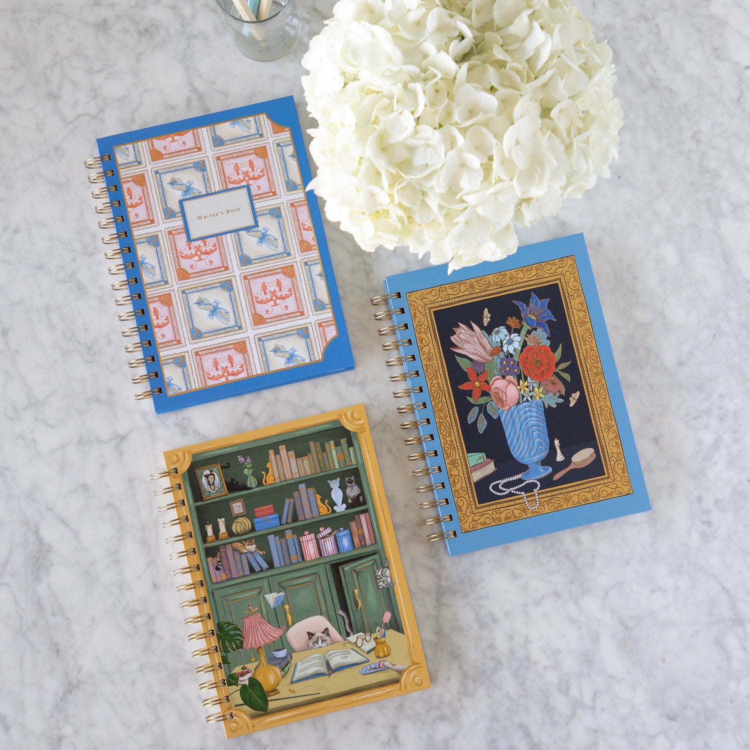 Assorted spiral notebooks by Good Juju Ink next to a vase of flowers and a cup of pencils.