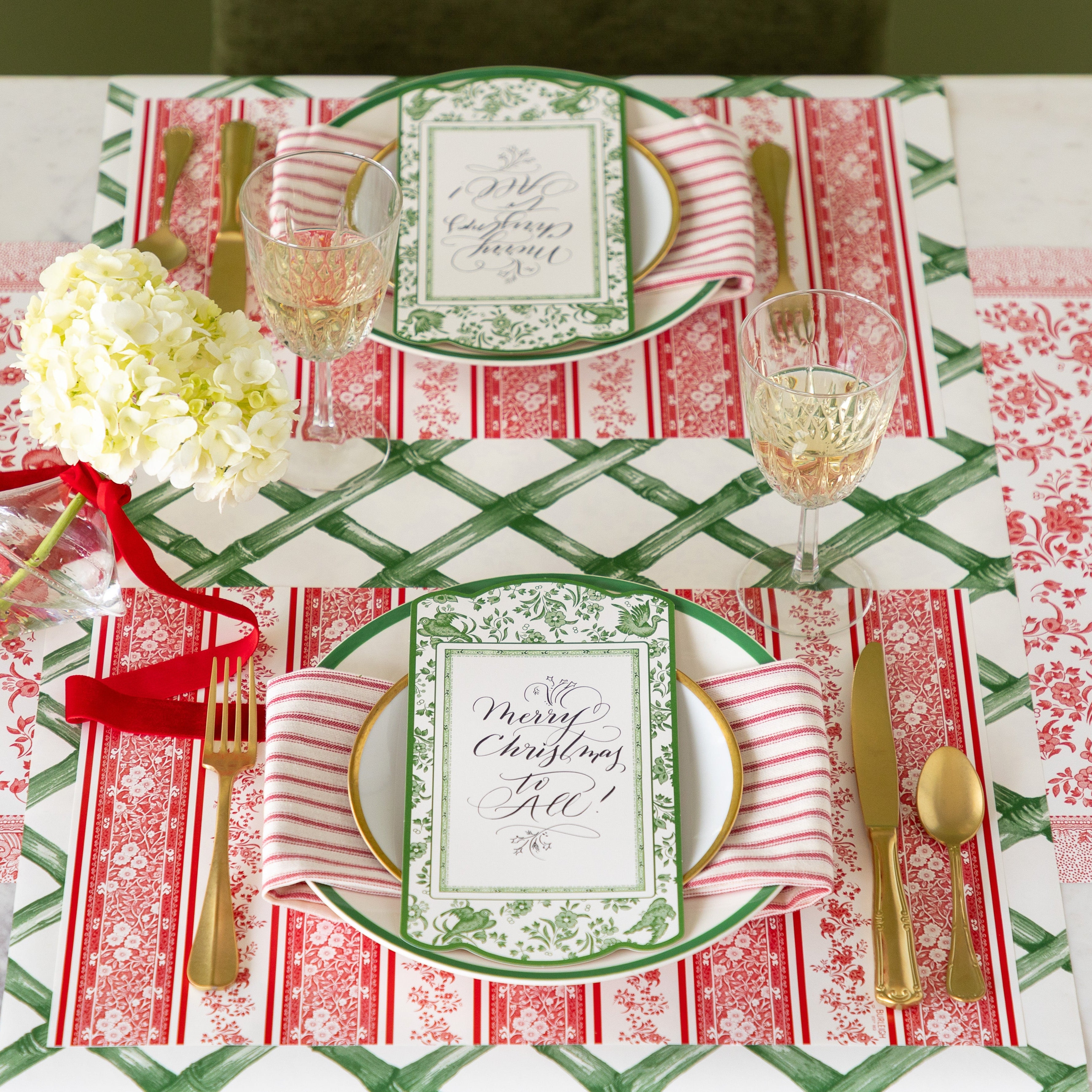 A table setting featuring the Red Burleigh Stripe Placemat, green rimmed dinner and gold rimmed salad plates with a red stripe napkin and Burleigh Green Regal Peacock Table Accent reading "Merry Christmas to All!" atop, gold flatware, glass of wine and Green Lattice Runner and Red Burleigh Runner underneath.