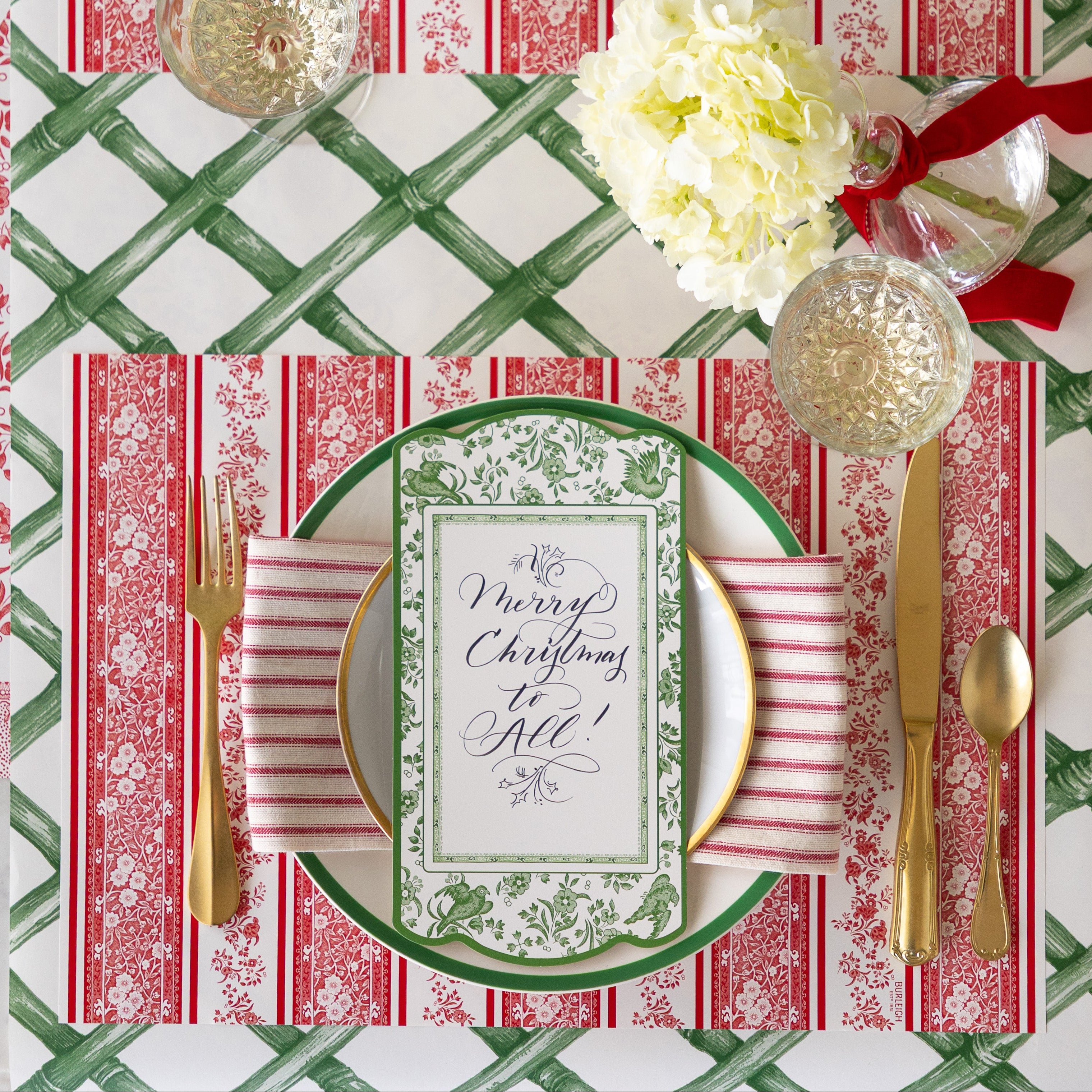 A place setting featuring the Red Burleigh Stripe Placemat, green rimmed and gold rimmed dinner plates with a red stripe napkin and Burleigh Green Regal Peacock Table Accent reading "Merry Christmas to All!" atop, gold flatware, glass of wine and Green Lattice Runner underneath.