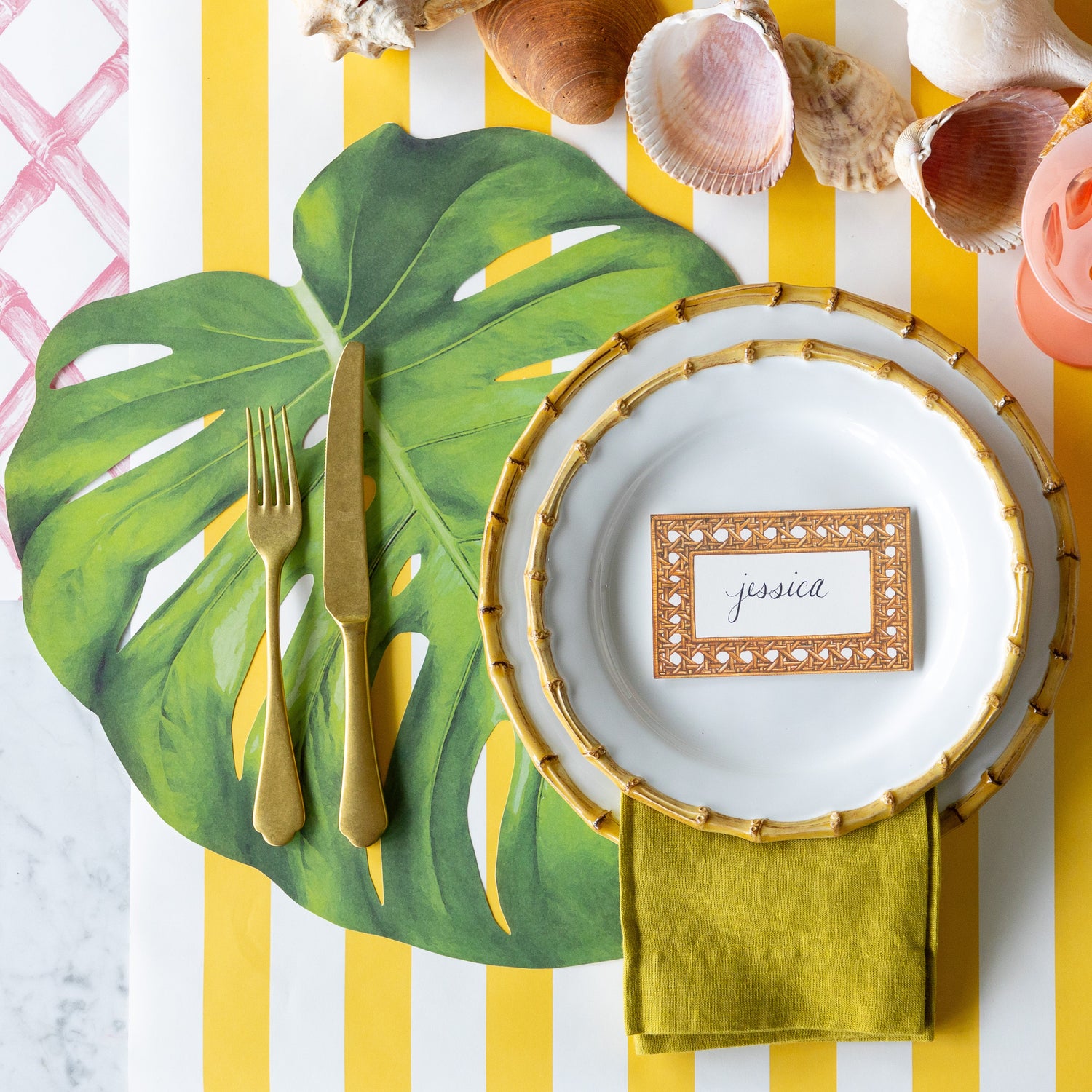 A tropical place setting featuring the Die-cut Monstera Leaf Placemat, bamboo rimmed dinner plates with the Rattan Weave Place Card reading &quot;jessica&quot; atop, seashells, gold flatware and a green napkin on the Marigold and Pink Lattice Runners.