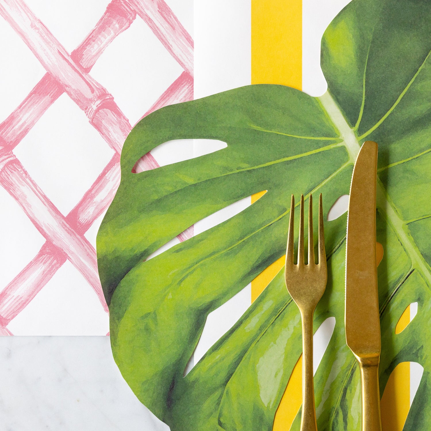 The Pink Lattice Runner under the Marigold Classic Stripe Runner and Monsterra Leaf Placemat with gold flatware atop.