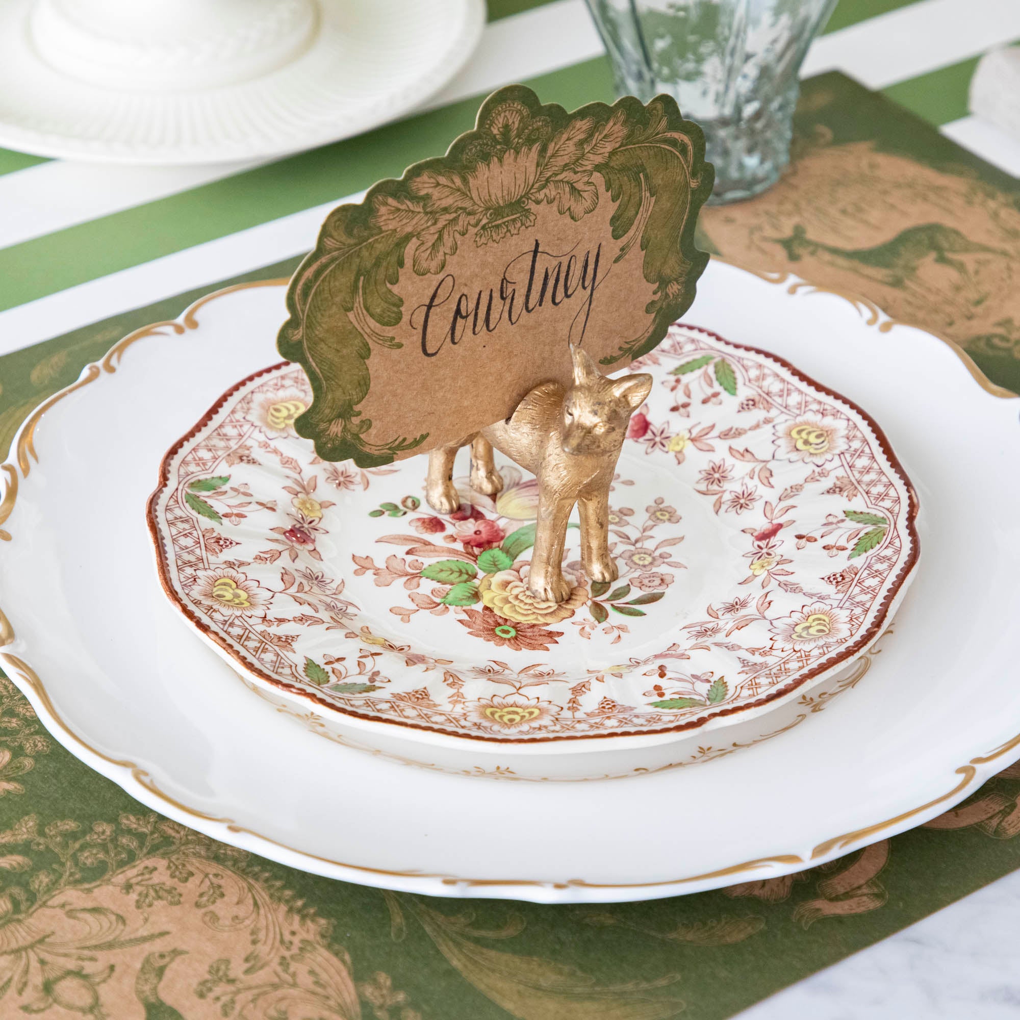 The Fox Name Card Holder holding the Moss Fable Toile Place Card with "Courtney" written in cursive, standing on two dinner plates with the Moss Fable Toile Placemat and Moss Green Classic Stripe Runner underneath.