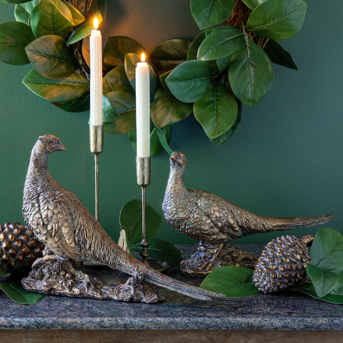 2 decorative bronze colored pheasants on a mantle with candlesticks and assorted greenery. 
