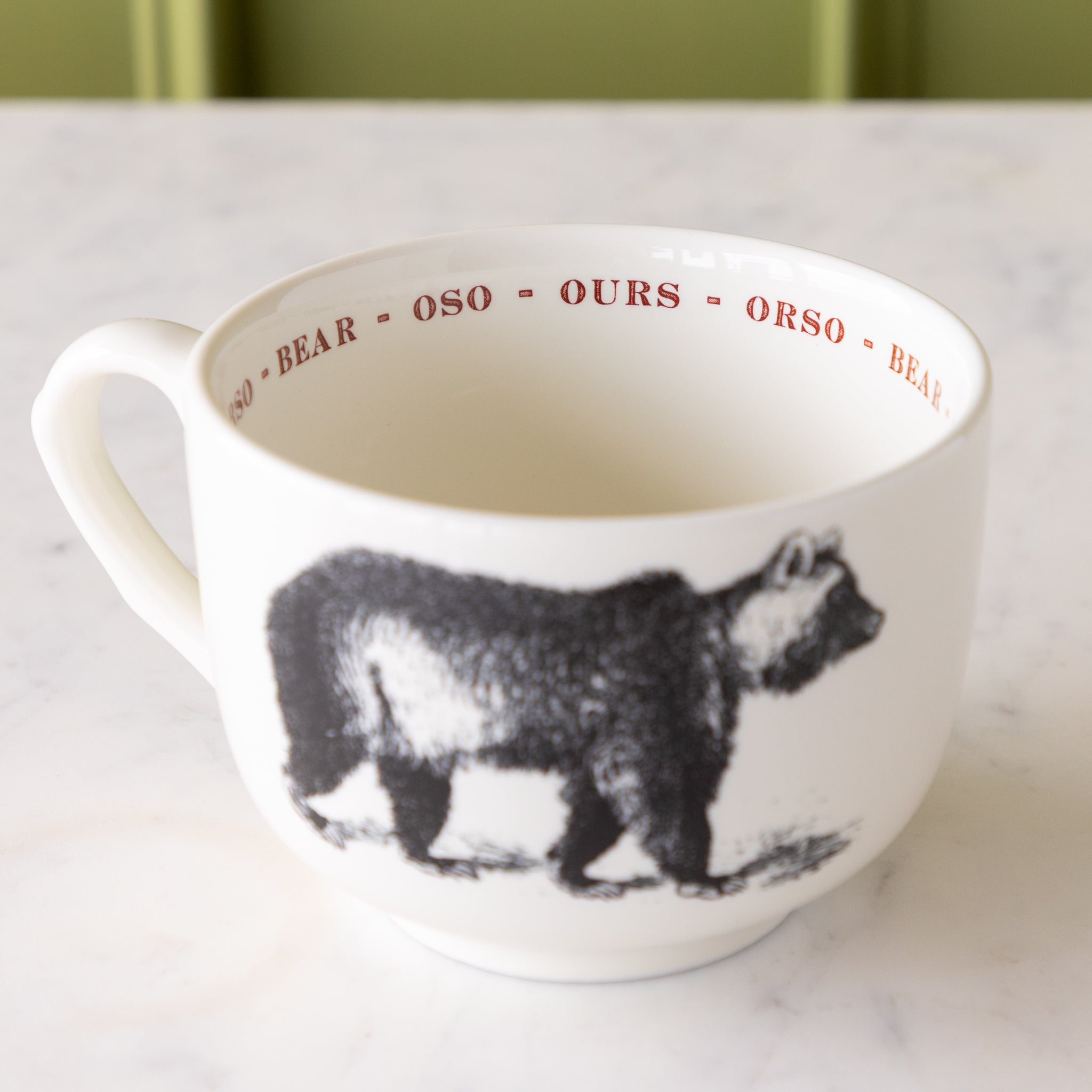White mug with black bear illustration with "bear" in different languages in red text around the inside rim.