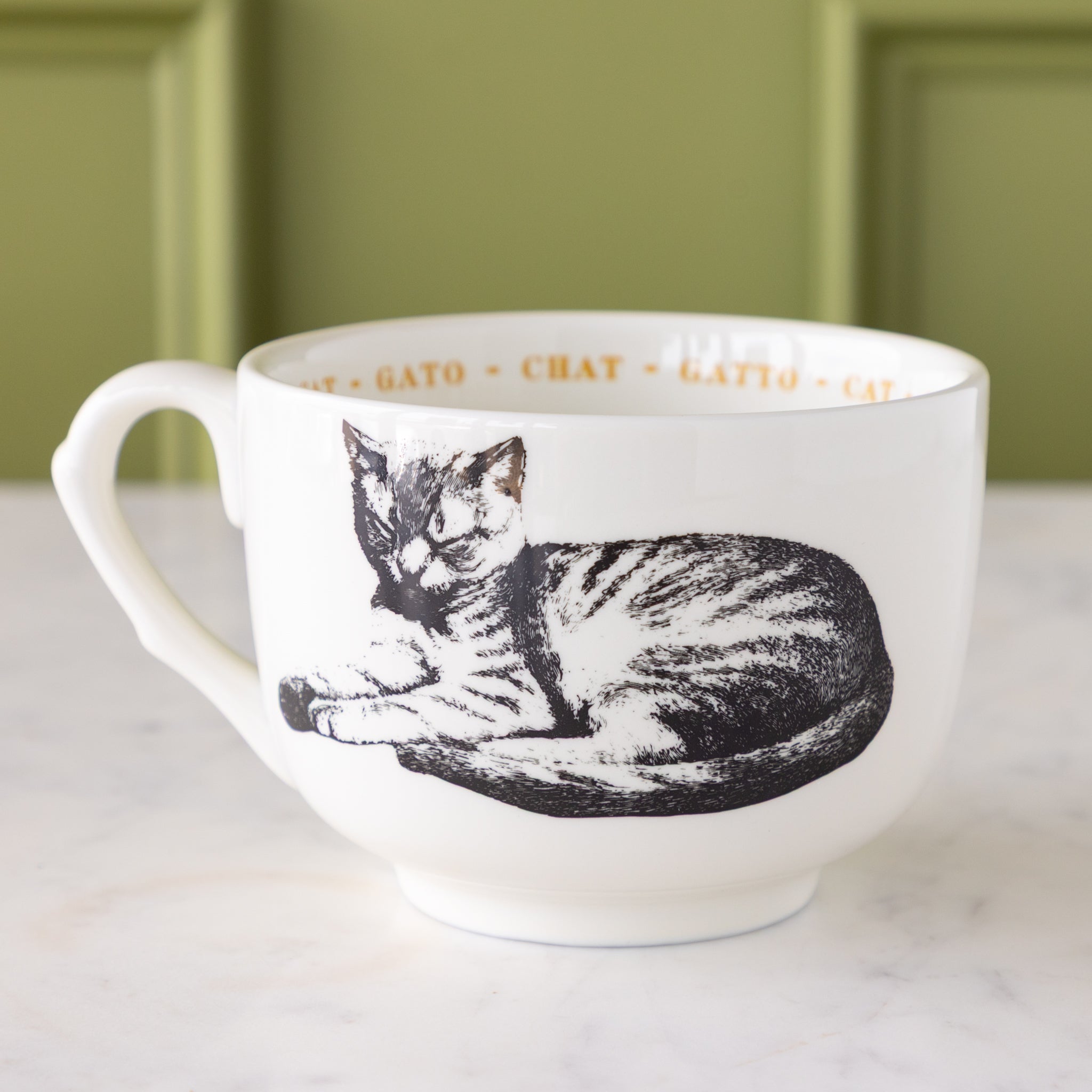 White mug with black cat illustration with "cat" in different languages in yellow text around the inside rim.