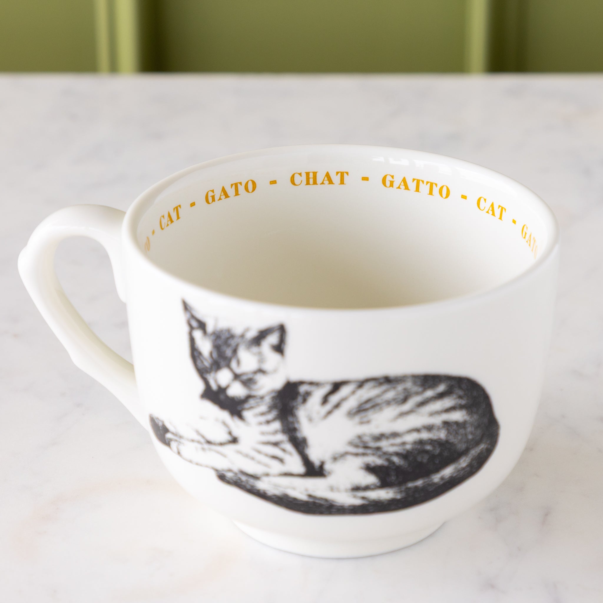 White mug with black cat illustration with "cat" in different languages in yellow text around the inside rim.