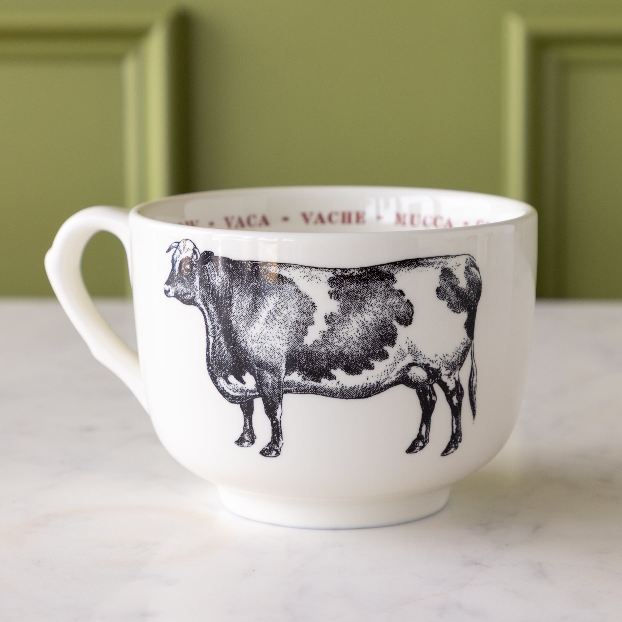 White mug with black cow illustration with "cow" in different languages in red text around the inside rim.