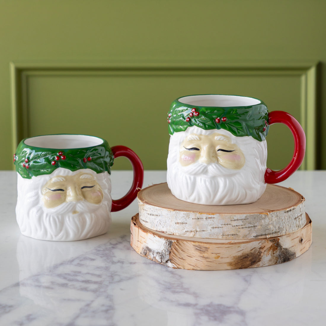 Two vintage Santa mugs with a sculpted design and hand-painted green and red details sit on a marble surface, one atop two wooden slices, against a green paneled background.