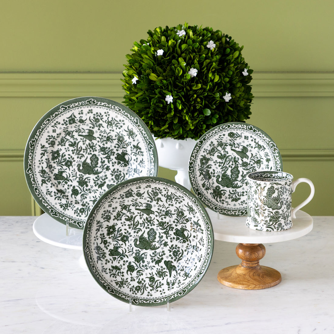 Burleigh Green Regal Peacock Dinnerware displayed on a table with topiary behind it.