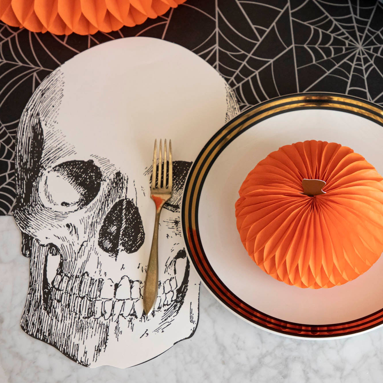 A spooky table setting featuring the Die-cut Skull Placemat, offset to the left of a place setting with a Honeycomb Pumpkin atop, and Spiderweb Runner underneath.