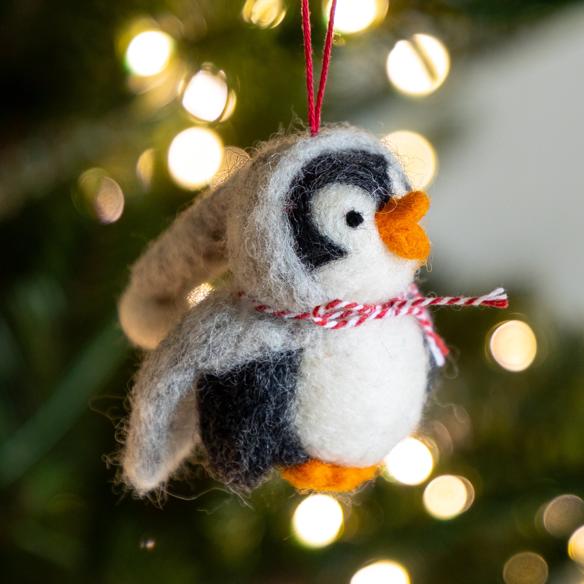 Felted penguin ornament with a red and white striped scarf against a blurred Christmas tree background.