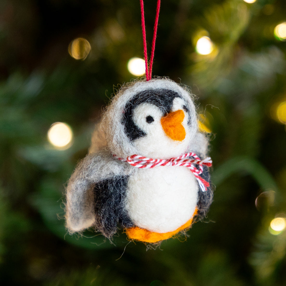 Felted penguin ornament with a red and white striped scarf against a blurred Christmas tree background.