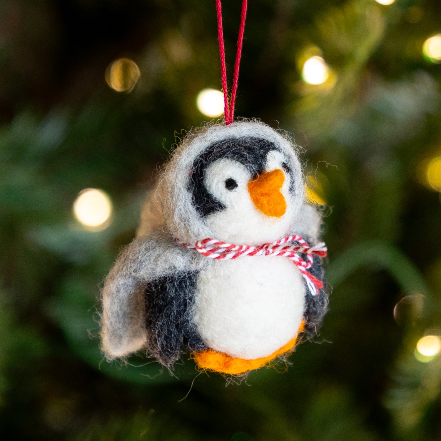 Felted penguin ornament with a red and white striped scarf against a blurred Christmas tree background.