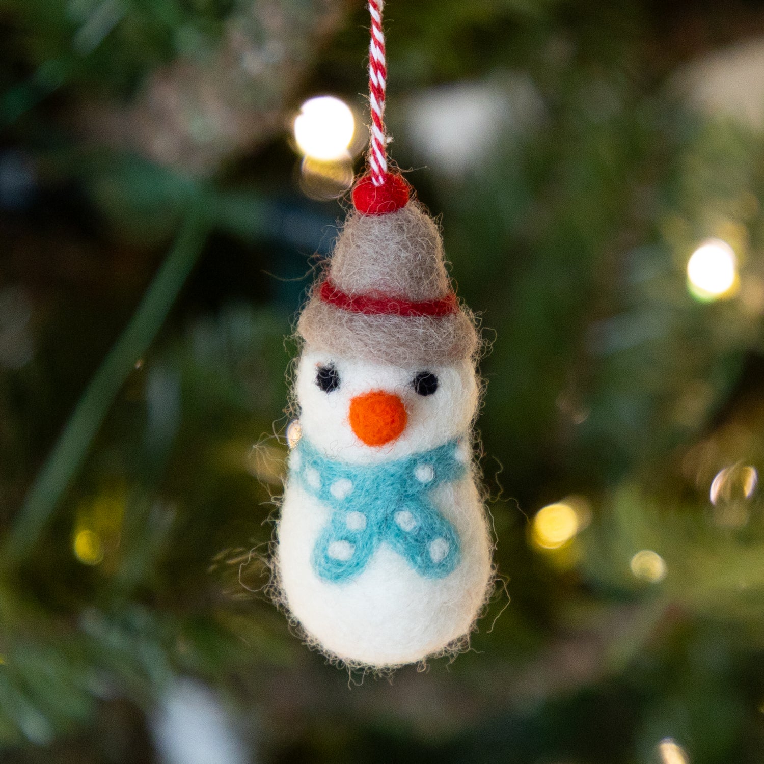 Felt snowman ornament hanging on a Christmas tree with blurred lights in the background.