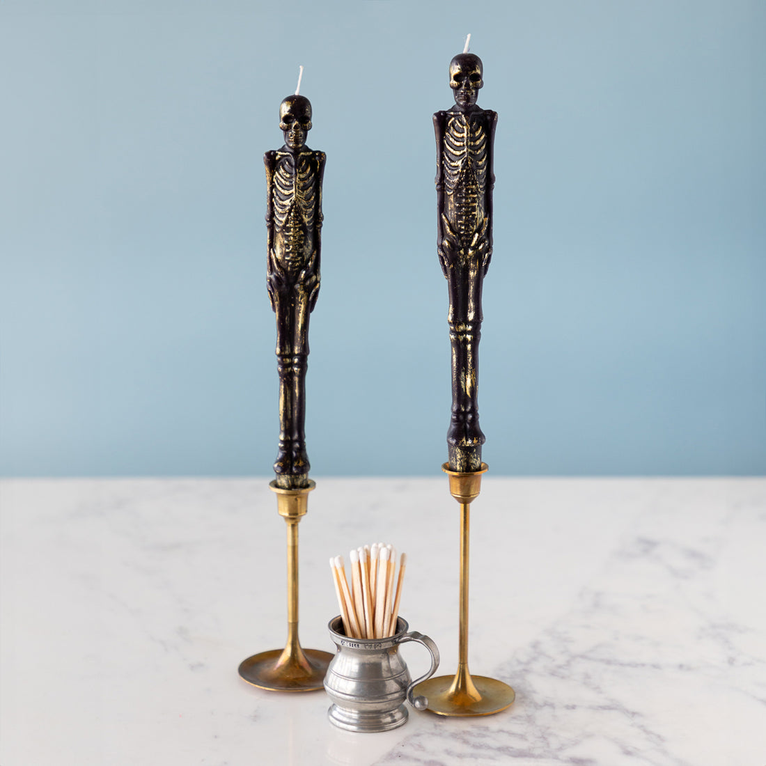 Two skeleton candles in gold candle holders next to a container with matches on a blue background.