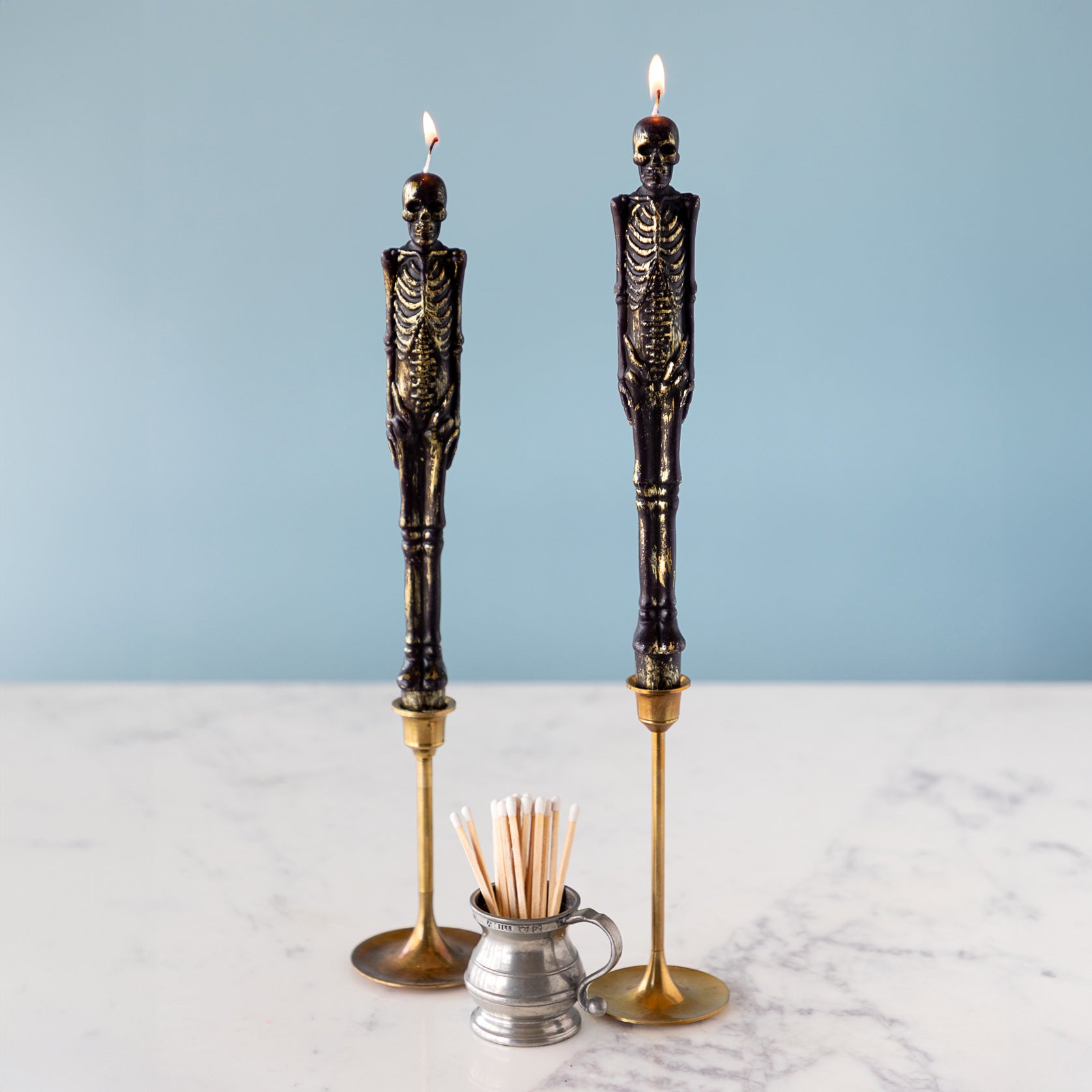 Two lit Skeleton Taper Candles in gold candle holders next to a container with matches on a light blue background.